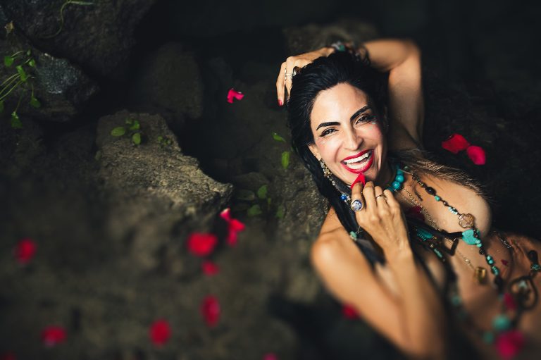 "Sol Tamargo, Erotic Godmother, sonríe entre pétalos de rosa y piedras — collares turquesa, ritual de sensualidad y erotismo sagrado"