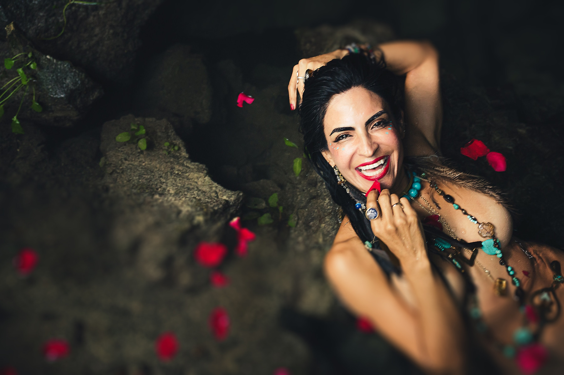 "Sol Tamargo, Erotic Godmother, sonríe entre pétalos de rosa y piedras — collares turquesa, ritual de sensualidad y erotismo sagrado"